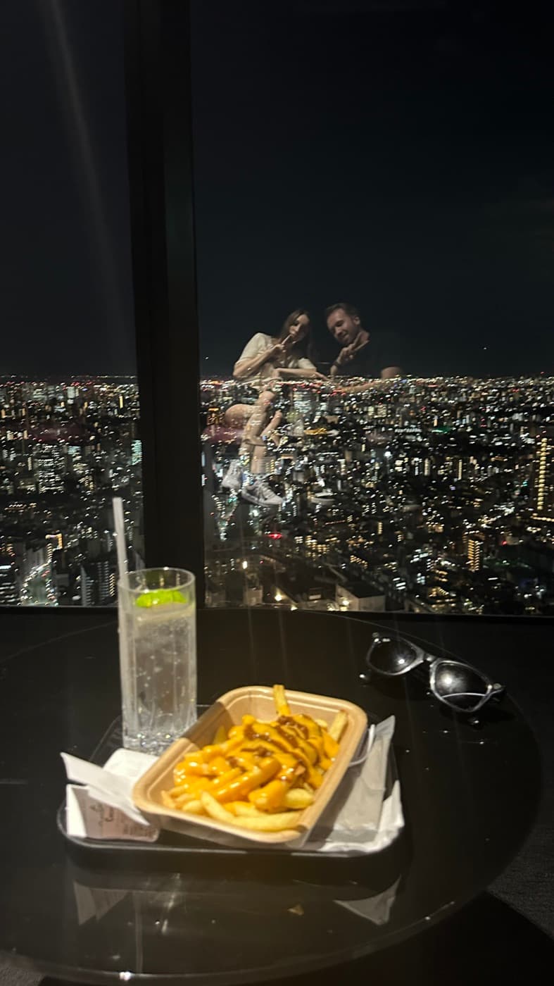 Tisch in der Bar mit Pommes und einem Longdrink, dahinter das nächtliche Lichtermeer hinter der Scheibe
